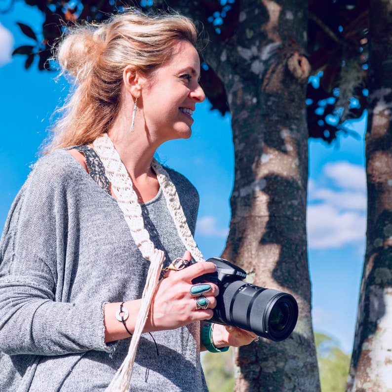 May include: A woman wearing a gray sweater and a white crocheted strap holds a black camera with a long lens. She is standing in front of a tree trunk.