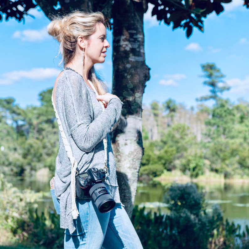 May include: A woman wearing a gray sweater and blue jeans is standing in front of a tree. She is holding a black camera with a lens attached to it. The camera is hanging from a white strap around her neck.