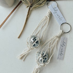 May include: Two macrame keychains with silver disco ball ornaments. Each keychain has a white tag with the text "lily cords handmade macrame".