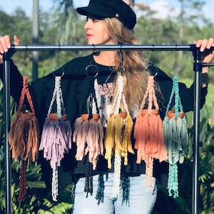 May include: A woman is holding up a rack of macrame plant hangers in various colors. The hangers are made with cotton rope and have a small terracotta pot at the bottom. The woman is wearing a black sweater and blue jeans.