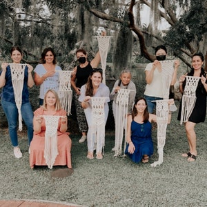 May include: A group of people holding up various macrame wall hangings. The macrame pieces are in different shapes and sizes, with a natural, off-white color. The background features a lush green lawn and trees.
