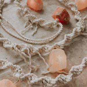 May include: A white macrame rope garland with pink Himalayan salt rocks strung throughout. The garland is on a light beige surface.