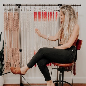 May include: A woman sits on a stool in front of a rack of macrame wall hangings. The wall hangings are made with natural colored cord and ombre red and white cord. The rack is labeled "Lily Cords".