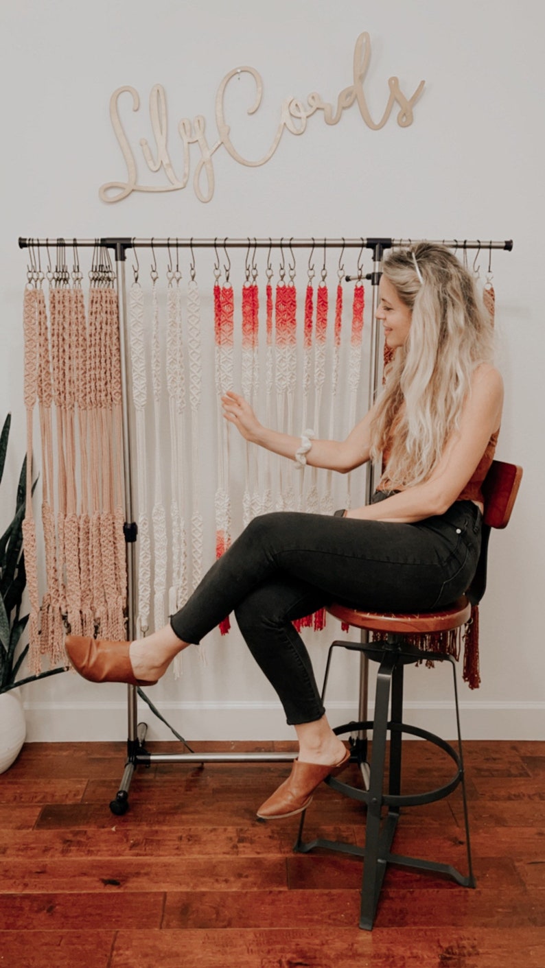 May include: A woman sits on a stool in front of a rack of macrame wall hangings. The wall hangings are made with natural colored cord and ombre red and white cord. The rack is labeled "Lily Cords".