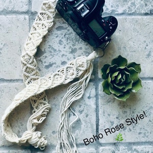 May include: A white macrame camera strap with long tassels. The strap is attached to a black DSLR camera. The text "Boho Rose Style!" is written in white on a gray background.