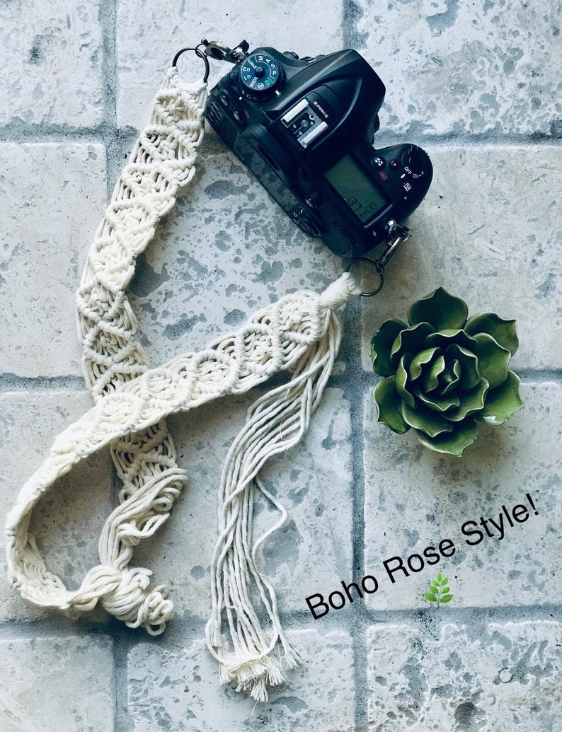 May include: A white macrame camera strap with long tassels. The strap is attached to a black DSLR camera. The text "Boho Rose Style!" is written in white on a gray background.