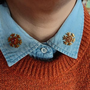Könnte beinhalten: Zwei goldfarbene Schneeflocken-Anstecknadeln sind an einem hellblauen Jeanshemdkragen befestigt. Die Anstecknadeln haben ein lächelndes Gesicht in der Mitte und sind auf einem orangefarbenen Pullover angebracht. Die Anstecknadeln verleihen dem Outfit eine festliche Note.