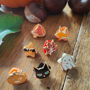 Puede incluir: Una colección de broches de esmalte con temas de otoño y Halloween. Los broches incluyen una calabaza, un murciélago, un fantasma, un gato negro, un zorro y una hoja de arce. Los broches están colocados sobre una superficie de madera con castañas y una calabaza en el fondo.