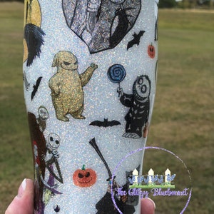 May include: White glitter tumbler with a Nightmare Before Christmas theme. The tumbler features Jack Skellington, Sally, Oogie Boogie, and other characters from the movie. The tumbler is decorated with black and white illustrations and a glittery finish.