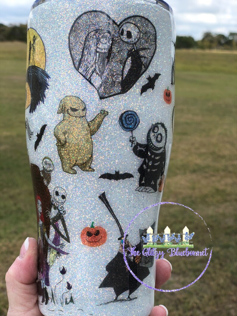 May include: White glitter tumbler with a Nightmare Before Christmas theme. The tumbler features Jack Skellington, Sally, Oogie Boogie, and other characters from the movie. The tumbler is decorated with black and white illustrations and a glittery finish.