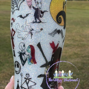 May include: A white glitter tumbler with a variety of characters from the Nightmare Before Christmas movie. The characters include Jack Skellington, Sally, Oogie Boogie, and more. The tumbler has a black and white spider web design and a yellow moon with a silhouette of the characters.