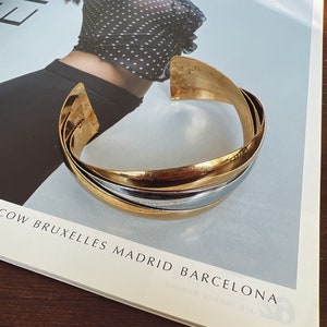 May include: A gold and silver bangle bracelet with a wide, curved design. The bracelet is made of metal and has a polished finish.