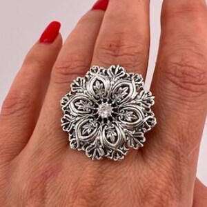 Könnte beinhalten: Ein großer, verzierter Silberring mit floralem Design. Der Ring hat einen zentralen, klaren Edelstein und aufwendige Details im gesamten blütenförmigen Design. Der Ring wird an einer Hand mit rotem Nagellack getragen.