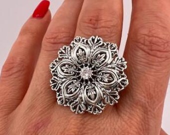 May include: A large, ornate silver ring with a floral design. The ring features a central, clear gemstone and intricate detailing throughout the flower-shaped setting. The ring is worn on a hand with red nail polish.