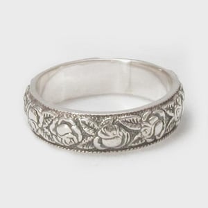 Può includere: Un anello d'argento con un motivo floreale. L'anello presenta un motivo ripetuto di rose e foglie attorno alla fascia. L'anello è realizzato in un materiale metallico lucido ed è posto su uno sfondo bianco.