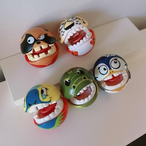 Peut inclure: Six balles en plastique colorées avec des visages d'animaux et des bouches ouvertes avec des dents. Les animaux sont un raton laveur, un guépard, un pingouin, un alligator et deux autres animaux.