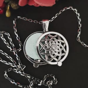 Può includere: Collana con pendente argentato con un design traforato circolare. Il pendente presenta un motivo floreale centrale con una piccola gemma e un vano incernierato contenente un inserto verde chiaro. Anche la catena della collana è argentata.