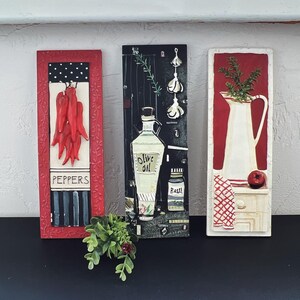 May include: Three framed wall art pieces with a kitchen theme. The first piece features red peppers, the second piece features a bottle of olive oil and a jar of basil, and the third piece features a white pitcher and a red apple.