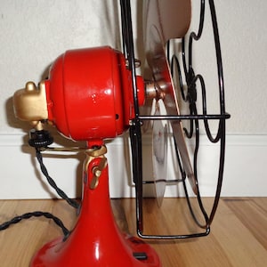 May include: A vintage electric fan in red and gold, featuring a black cage and blades. The fan has a red base and motor housing, with gold accents and a black power cord. The blades are a copper colour.
