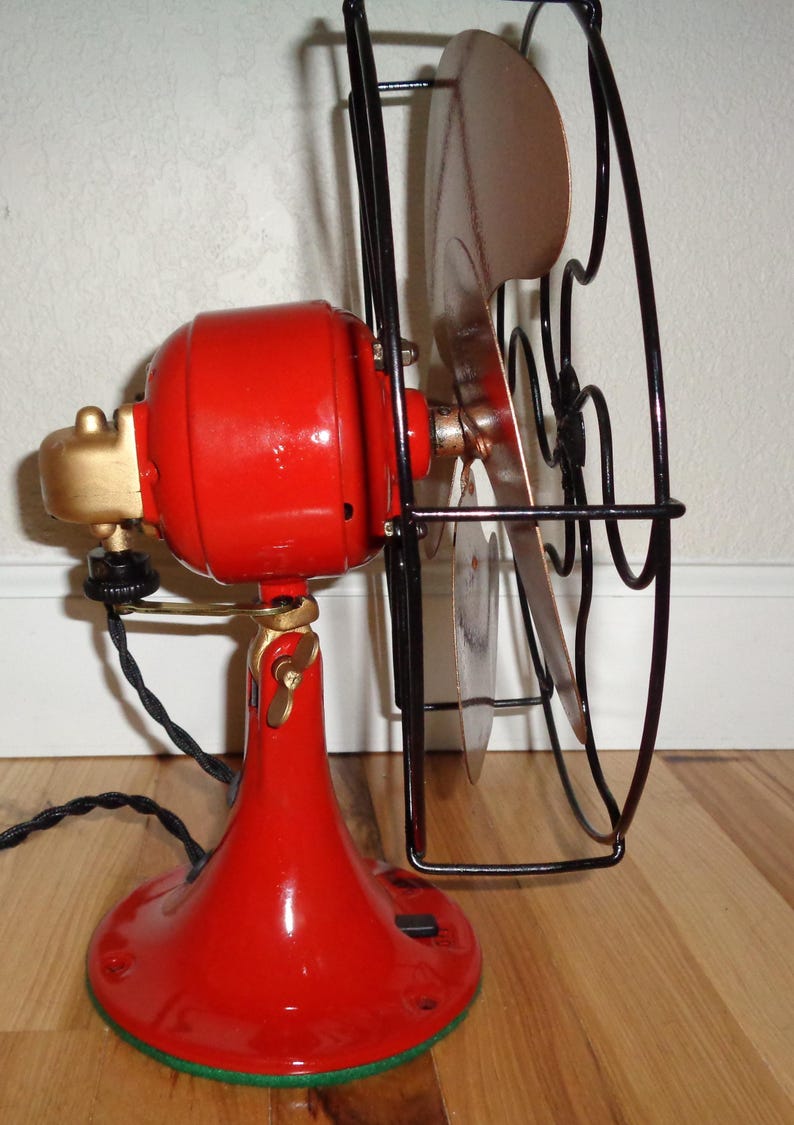 May include: A vintage electric fan in red and gold, featuring a black cage and blades. The fan has a red base and motor housing, with gold accents and a black power cord. The blades are a copper colour.