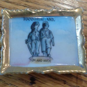 May include: A rectangular ceramic tray with a scalloped gold-painted rim. The centre features a blue and white illustration of two figures with the text HANNIBAL, MO. at the top and TOM AND HUCK printed at the bottom of the central graphic.