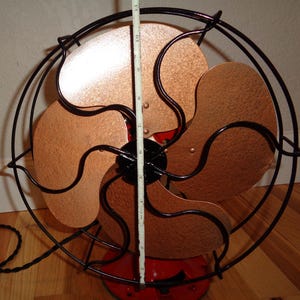 May include: An antique electric fan featuring copper-toned blades and a black wire guard. The fan blades measure approximately 25 cm in diameter. The base is red, and it has a black power cord.