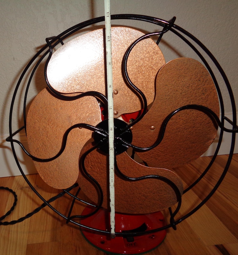 May include: An antique electric fan featuring copper-toned blades and a black wire guard. The fan blades measure approximately 25 cm in diameter. The base is red, and it has a black power cord.