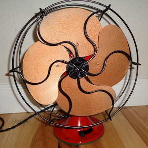 May include: An antique electric fan featuring copper-toned blades and a black wire guard. The fan has a red base and a black motor casing. The blades are arranged in a circular pattern. The fan is on a wooden floor.