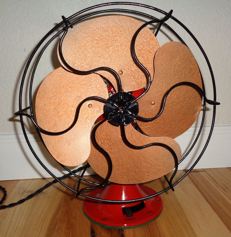 May include: An antique electric fan featuring copper-toned blades and a black wire guard. The fan has a red base and a black motor casing. The blades are arranged in a circular pattern. The fan is on a wooden floor.