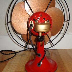 May include: A vintage electric fan featuring a red base and motor housing, gold detailing, and copper-toned blades. The fan is encased in a black wire guard with a black power cord. A classic home decor item.