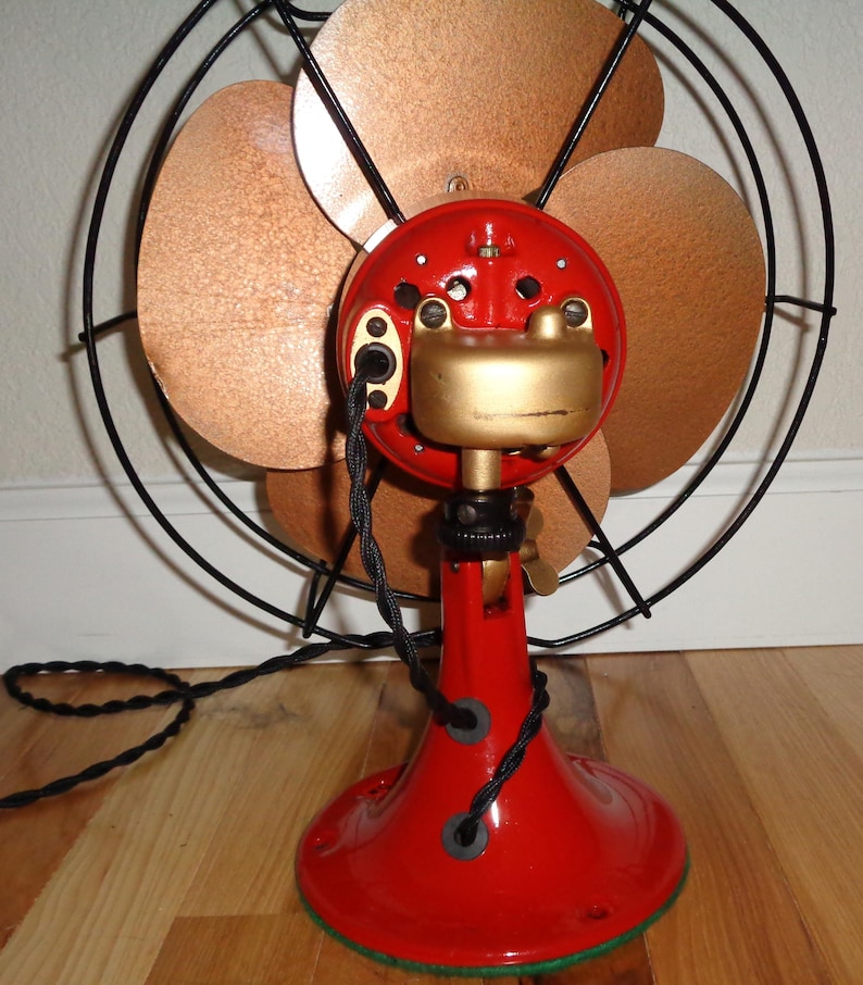 May include: A vintage electric fan featuring a red base and motor housing, gold detailing, and copper-toned blades. The fan is encased in a black wire guard with a black power cord. A classic home decor item.
