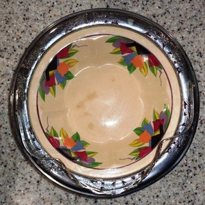 May include: A vintage cream-colored ceramic bowl with a silver rim and a floral design in shades of red, blue, yellow, and green. The bowl is decorated with stylized flowers and geometric shapes.
