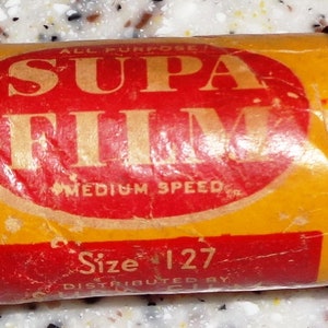 May include: A yellow and red film canister with the text "Supa Film" in a red circle and "All Purpose Medium Speed" in smaller text below the circle. The text "Size 127" is printed in red on the bottom of the canister.