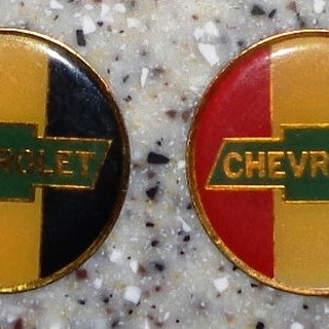 May include: Two gold-colored metal pins with a red, yellow, and black striped design. The center of each pin features a green and gold Chevrolet logo.