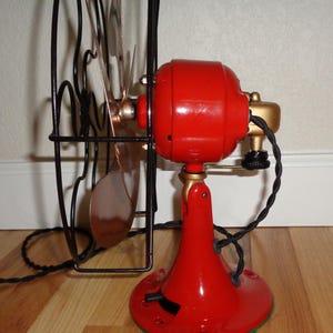 May include: A vintage red electric fan with a black cage and copper-coloured blades. The fan has a gold-coloured motor housing and base, and a black power cord. The fan is on a wooden floor.