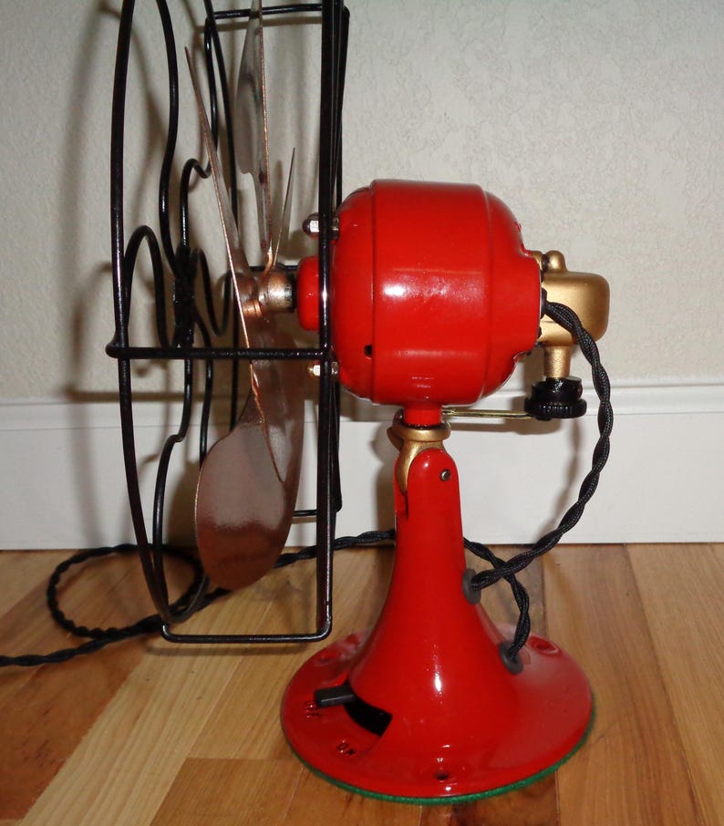 May include: A vintage red electric fan with a black cage and copper-coloured blades. The fan has a gold-coloured motor housing and base, and a black power cord. The fan is on a wooden floor.