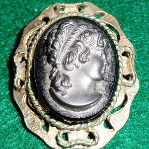 Può includere: Spilla cammeo vintage di forma ovale con il profilo di una donna con un copricapo di piume. Il cammeo nero è incastonato in una cornice di metallo dorato con un bordo a forma di corda e dettagli decorativi in filigrana. Un gioiello classico.