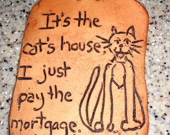 May include: A rectangular, terracotta-colored plaque with a cat illustration and the text "It's the cat's house, I just pay the mortgage." The plaque has a dark brown outline and a loop for hanging.