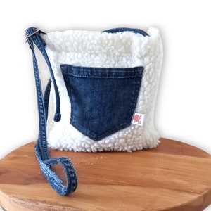 Op de afbeelding: Een witte nep bont crossbody tas met een blauwe denim zak en een blauwe denim band. De tas heeft een klein roze label met een wit ontwerp.
