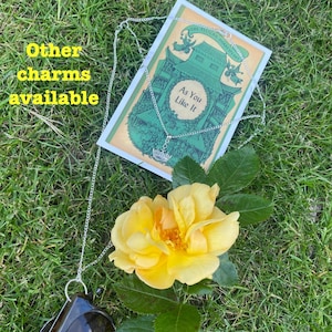 May include: A silver chain necklace with a crown charm, laying on a book titled "As You Like It" by William Shakespeare. The book is on a bed of green grass with a yellow rose.