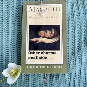 May include: A close-up of a book titled "Macbeth" by William Shakespeare. The book is open and a silver charm necklace with a crown pendant is hanging from the top of the book. The book is on a blue knitted surface.