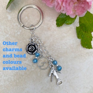 Op de afbeelding: Zilverkleurige sleutelhanger met een rozenbedel, blauwe kralen en een kurkentrekkersbedel. De tekst "Other charms and bead colours available" is in het blauw weergegeven. Roze bloemen zijn op de achtergrond.