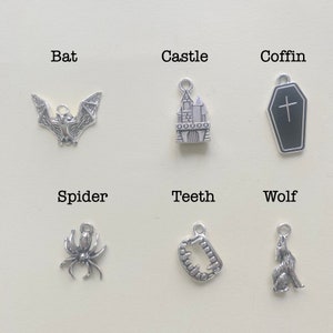 May include: Six silver charms with Halloween themes. The charms are bat, castle, coffin, spider, teeth, and wolf.