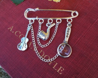 May include: A silver safety pin brooch with a chain and charms. The charms include a magnifying glass, a pipe, and a guitar. The brooch is on a red book with the text "A.C. DOYLE" visible.
