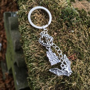 May include: Silver keychain with a bat, a small house, and a set of fangs hanging from a silver ring. The keychain is on a green mossy surface.