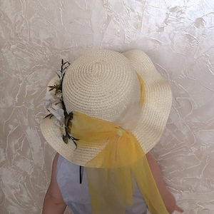 May include: A white straw hat with a yellow ribbon and white flowers. The hat has a wide brim and is decorated with a yellow tulle bow.