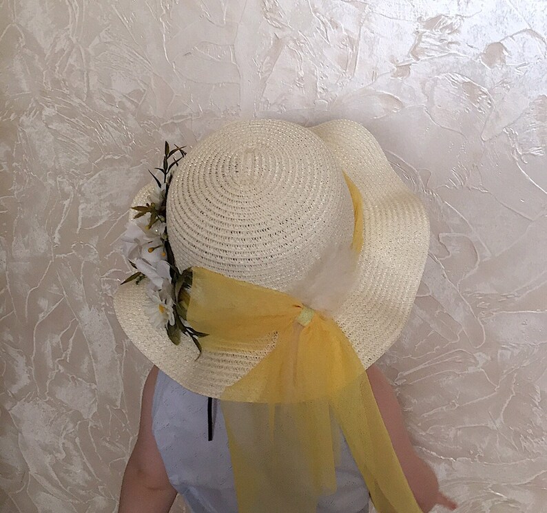 May include: A white straw hat with a yellow ribbon and white flowers. The hat has a wide brim and is decorated with a yellow tulle bow.