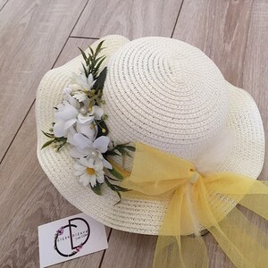 May include: A white straw hat with a yellow ribbon and a white flower embellishment. The hat has a wide brim and is decorated with a yellow tulle bow.