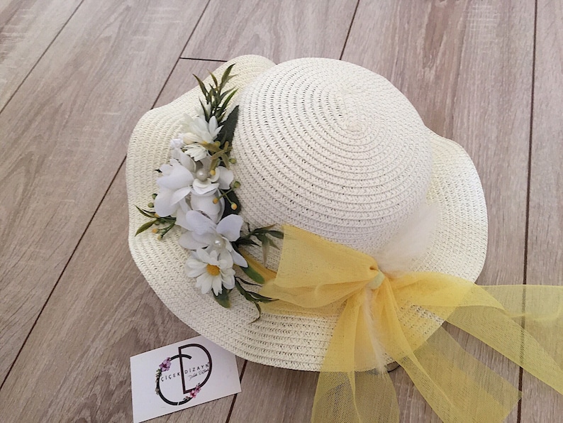 May include: A white straw hat with a yellow ribbon and a white flower embellishment. The hat has a wide brim and is decorated with a yellow tulle bow.
