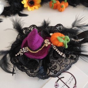 May include: A Halloween-themed hair accessory featuring a purple witch hat, an orange pumpkin, and black feathers. The base is black lace, embellished with rhinestones and a gold chain. Additional Halloween decorations are visible.